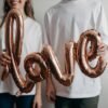A couple in white t-shirts holding a metallic 'love' balloon, expressing joy and affection.